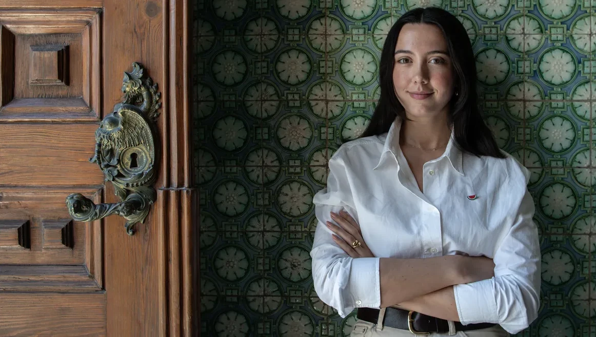 Foto de biografia da Leonor Caldeira. A Leonor aparece a meio corpo, a olhar directamente para a camera, com um sorriso confiante e um blazer branco, contrea um fundo vermelho.